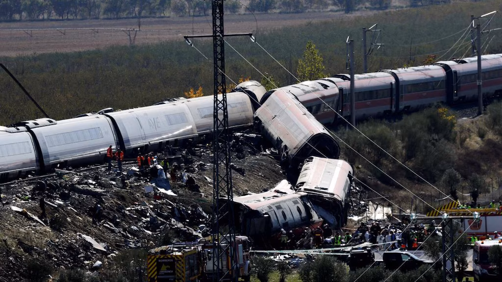 Train carriages leaning on the side, some spilling from the railway after a violent collision between two trains in the Spanish countryside. Rescue personnel is milling around, with ambulances and fire trucks parked next to the path.