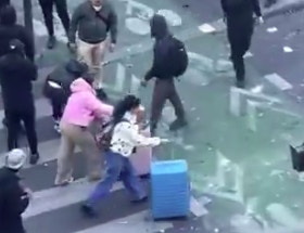 两名身穿粉嫩系服饰的女游客拖著行李箱,在一整群身穿黑衣的抗议人群中显得格外醒目。(X@RafaelSereti)