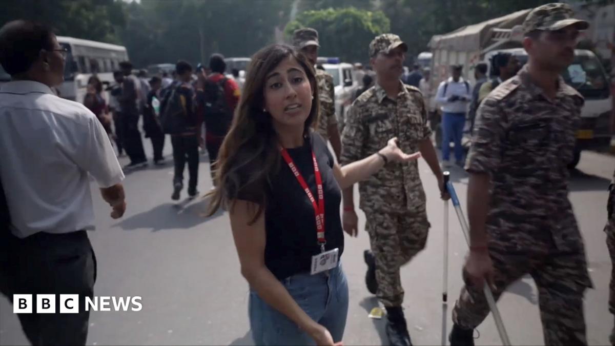 A female BBC correspondent wearing a t-shirt and jeans is seen pointing behind her as Indian security members walk past